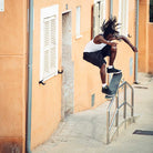 A skateboarder with long dreadlocks jumps over a handrail outside an orange building, wearing a white tank top, black shorts, and adidas Superskate x Kader Shoes JQ4413 in Core Black/Off White suede and leather.