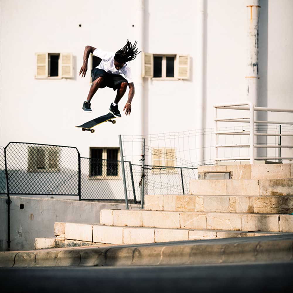 A skateboarder with dreadlocks does a mid-air trick above stone steps, wearing Superskate x Kader Shoes JQ4413 in core black and off white leather/suede from adidas Skateboarding, set against an urban scene with a white building and windows.