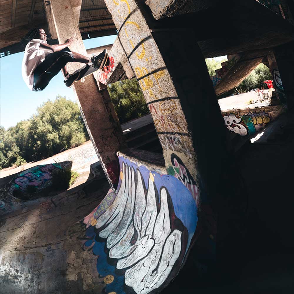 A skateboarder in Superskate x Kader Shoes JQ4413 Core Black Off White Leather Suede Adidas Skateboarding Shoes does a wall ride on a graffiti-covered pillar in a sunlit, abandoned skatepark with concrete ramps and overgrown trees.