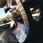 A skateboarder in Superskate x Kader Shoes JQ4413 Core Black Off White Leather Suede Adidas Skateboarding Shoes does a wall ride on a graffiti-covered pillar in a sunlit, abandoned skatepark with concrete ramps and overgrown trees.