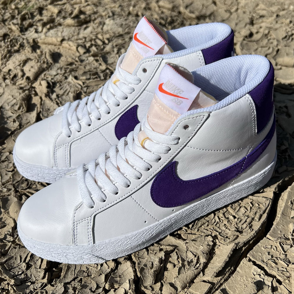 A pair of DZ4949-100 Court Purple White Blazer Mid Nike SB leather skateboarding shoes with purple swooshes and orange tongue accents, shown on rough textured ground.