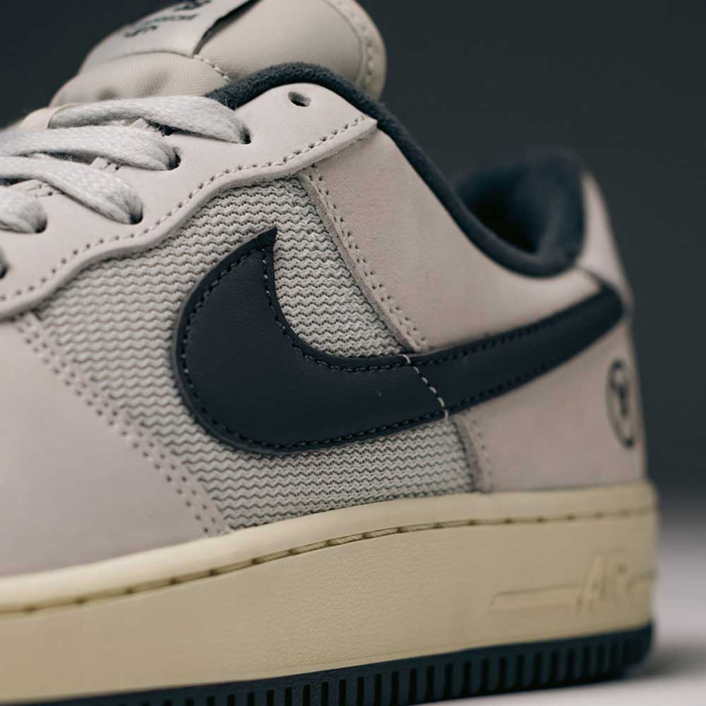 Close-up of the Yuto AF1 Iron Grey White Coconut Milk IO8439100 Nike SB Suede Skateboarding Shoe, highlighting the textured iron grey suede side panel, black Nike Swoosh logo, laces, and part of the coconut milk-toned rubber sole with signature Nike Air cushioning.