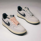 A pair of Yuto AF1 Iron Grey White Coconut Milk IO8439100 Nike SB Suede Skateboarding Shoes with black swooshes; the left shoe features pink laces, and the right shoe has white laces. Designed as premium skate shoes, both have dark soles, Nike Air cushioning for enhanced comfort, and are presented on a light background.