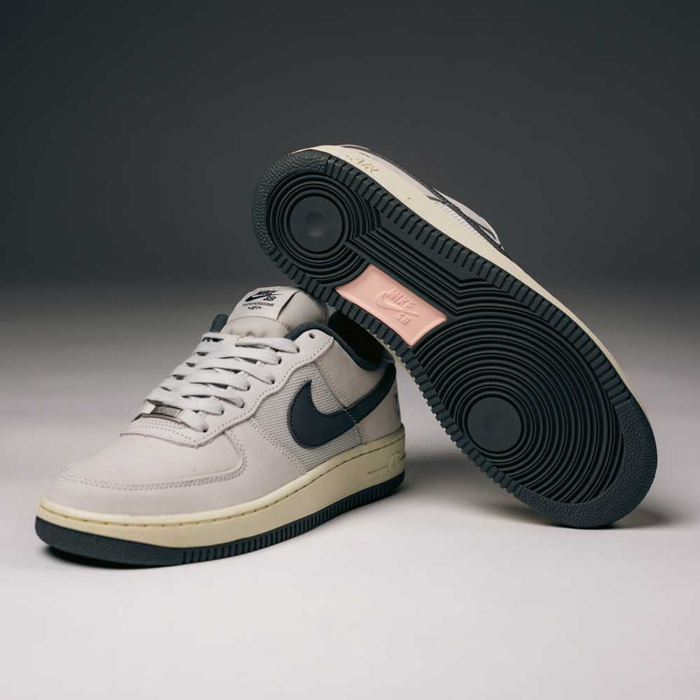 A pair of Yuto AF1 Iron Grey White Coconut Milk IO8439100 Nike SB Suede Skateboarding Shoes by Nike is displayed; one shoe stands upright to reveal the sole, while the other rests flat. These skate shoes feature classic Nike Air cushioning and are showcased against a softly lit, plain background.